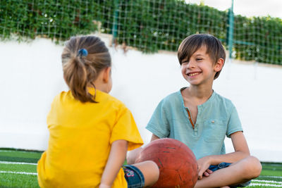 Smiling boy sitting on lawn with sister and talking with her and looking each other