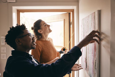 Couple assisting each other while hanging painting on wall near doorway at home