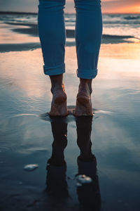 Low section of woman standing on sea shore against sky during sunset