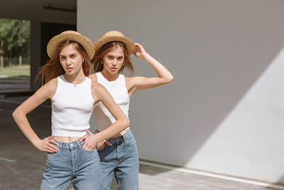 Twin sisters in white t-shirts, blue jeans and straw hats posing in urban area on a sunny day.