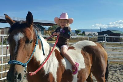 Full length portrait of happy girl sitting on horse at pen against sky