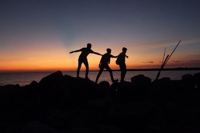 Silhouette friends on rocks by sea against sky during sunset