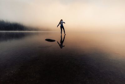 Silhouette of  young woman touching the water with legs in the foggy lake during autumn at sunrise.