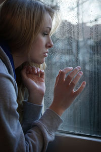 Thoughtful young woman by wet window at home during rainy season