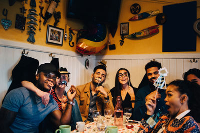 Portrait of cheerful young multi-ethnic friends holding props while sitting at restaurant during dinner party