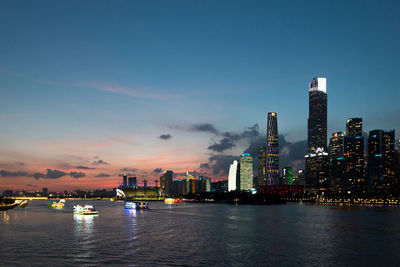 Illuminated buildings by river against sky during sunset