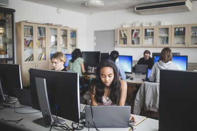 Female high school student using laptop at desk against teacher and friends sitting in computer lab