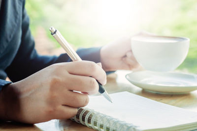 Midsection of man having coffee while writing on spiral notebook at table in cafe