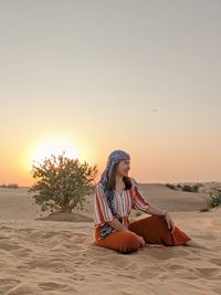 Woman sitting on sand in desert against clear sky during sunset