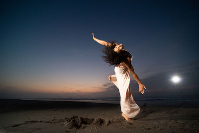 Full length of woman with arms raised standing at beach against sky during sunset
