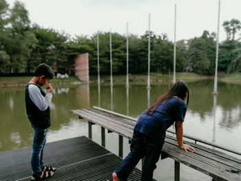 Boy and girl standing on footbridge over lake against sky