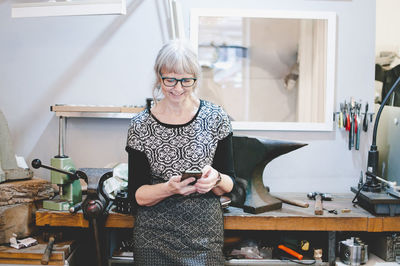 Smiling senior woman using smart phone in jewelry workshop