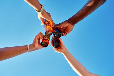 From below of excited unrecognizable young diverse best friends toasting with bottles of beer against cloudless blue sky on sunny summer day