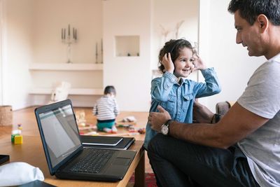 Smiling daughter with headphones standing by father while brother playing in background at home