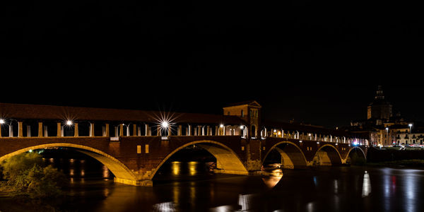 Illuminated bridge over river by buildings against sky at night in pavia, italy