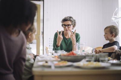 Senior female teacher communicating with students through hand signs while sitting at dining table during lunch break