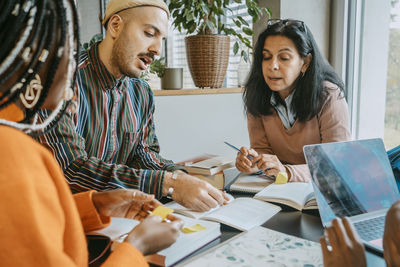 Multiracial students discussing over book while studying together at college cafeteria