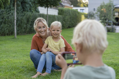 Boy photographing smiling mother and sister through smart phone in garden