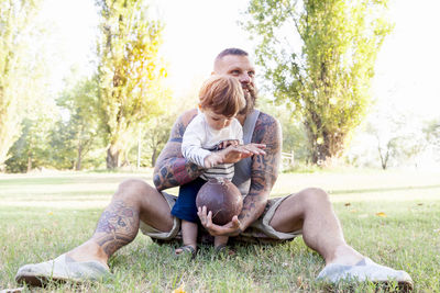 Smiling bearded man with son holding american football ball while sitting on grassy field