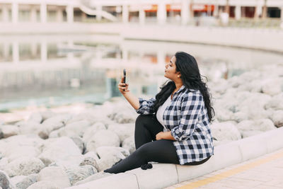 An indian girl takes a selfie on her phone while sitting on the embankment while walking in the city