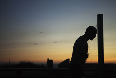 Silhouette man taking shower at beach against sky during sunset