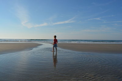 Full length rear view of boy standing in water at beach against sky