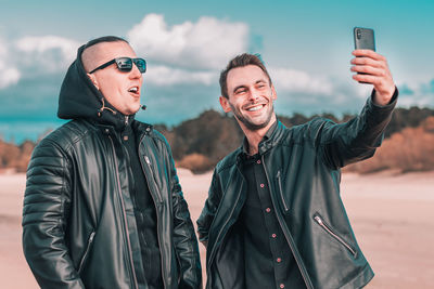 Smiling man taking selfie with male friend at beach against sky