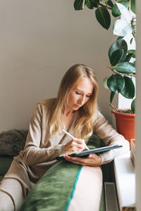 Beautiful blonde young woman artist illustrator drawing on tablet near window at home