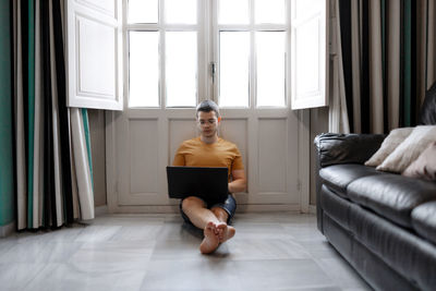 Focused male freelancer in casual outfit sitting on floor near window and typing on laptop while working remotely from home