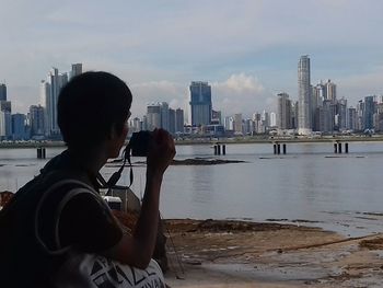 Man photographing sea with cityscape in background