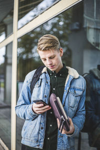 Young man using mobile phone while leaning on glass wall