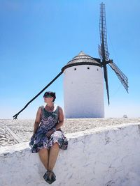 Woman wearing sunglasses while sitting on stone wall with traditional windmill in background