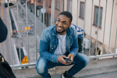 High angle view of smiling man crouching against railing while looking away on footbridge in city