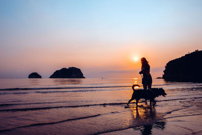 Silhouette mid adult woman with dog standing at beach against sky during sunset