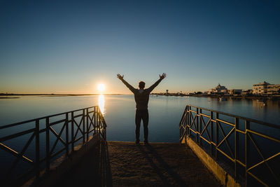 Rear view of man with arms raised standing on pier by sea