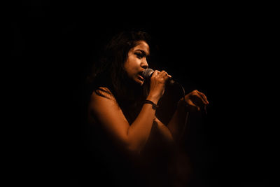 Side view of young woman singing on microphone against black background