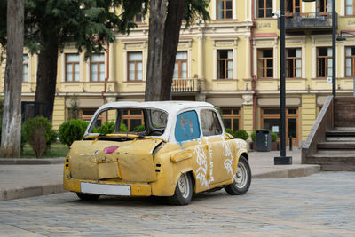 Broken down, old retro yellow white car park at sidewalk in city with house and trees in background.