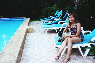 Portrait of smiling woman with dog sitting on lounge chair by swimming pool