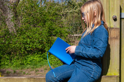 Girl enjoying reading from blue textbook while sitting on wooden fence.