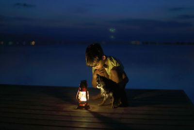 Child sitting with her dog at illuminated shore against sky at night
