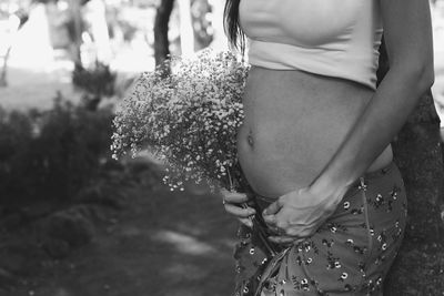 Midsection of pregnant woman holding flower standing by tree trunk