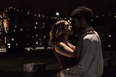 Side view of young couple standing against illuminated city at night