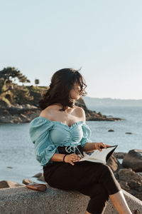 Woman reading book sitting against sea