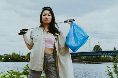 Low angle portrait of confident young woman picking garbage near river