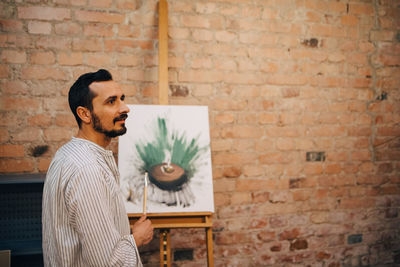 Side view of male artist standing by painting against brick wall at studio