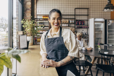 Portrait of happy female owner with tattooed arm standing near table in cafe