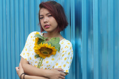 Portrait of woman holding sunflowers while leaning on corrugated iron