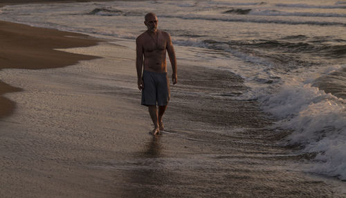 Rear view of shirtless man walking on beach during sunrise
