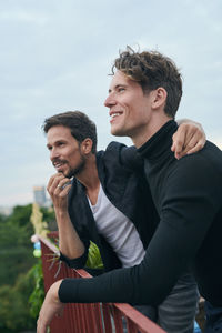 Smiling male friends looking away while standing by railing on terrace against sky