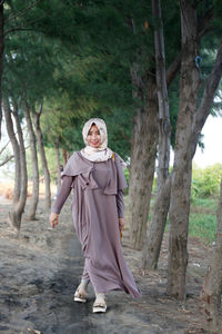 Full length portrait of smiling woman in hijab walking amidst trees